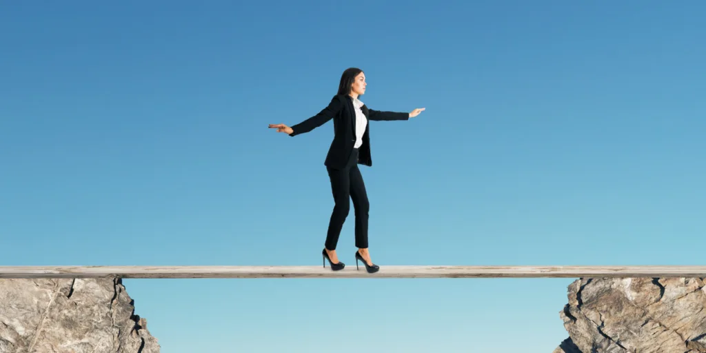 A woman in a business suit balancing on a narrow beam between two rocky cliffs, representing the challenges of navigating business decisions