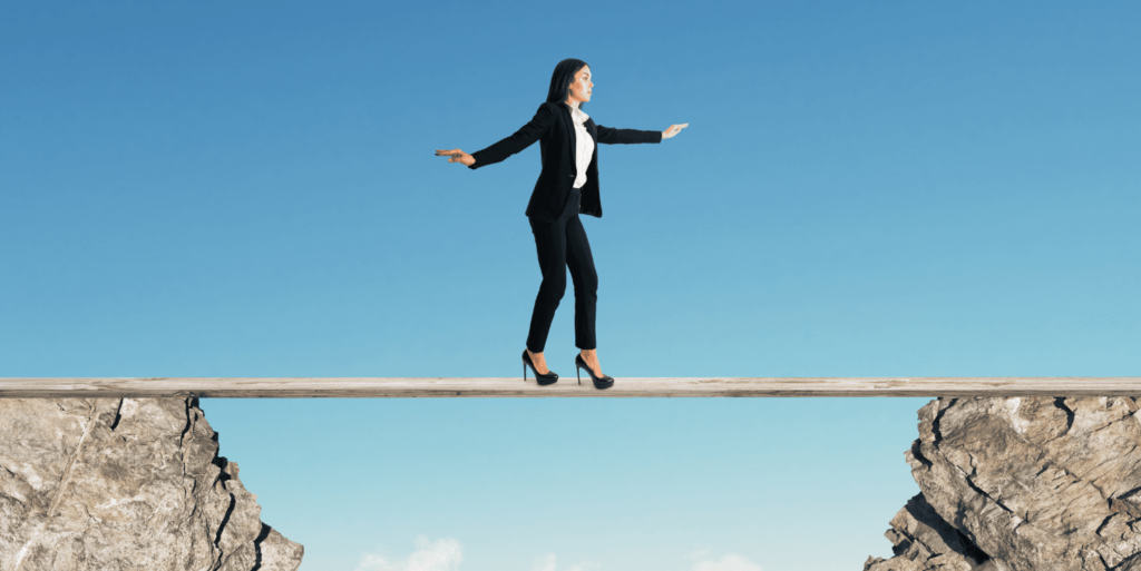 A woman in a business suit balancing on a narrow beam between two rocky cliffs, representing the challenges of navigating business decisions