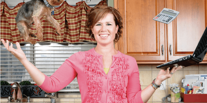 A woman in a pink cardigan smiles as she juggles kitchen items, including a frying pan, a laptop, and a small dog, illustrating multitasking in a lively kitchen setting