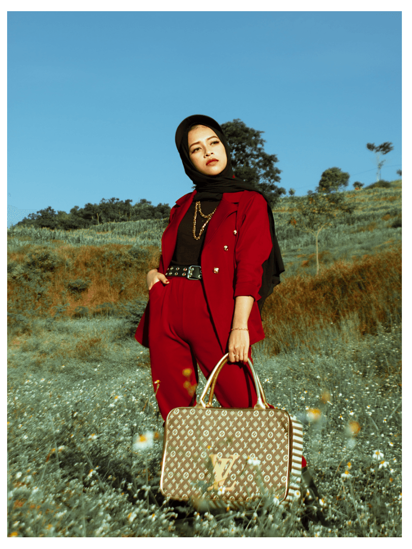 A fashionable woman wearing a stylish red suit and black hijab stands confidently in a green field. She holds a luxurious Louis Vuitton handbag, showcasing her sophisticated style against a backdrop of blue sky and rolling hills. The image emphasizes elegance and modern fashion.