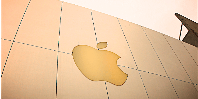 A close-up image of the Apple logo displayed prominently on a smooth surface, with soft lighting enhancing its golden hue against a minimalist background. The image conveys a sense of modern design and brand identity associated with Apple products.