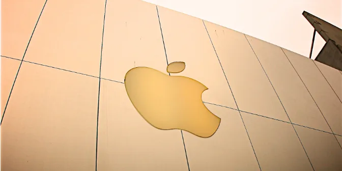 A close-up image of the Apple logo displayed prominently on a smooth surface, with soft lighting enhancing its golden hue against a minimalist background. The image conveys a sense of modern design and brand identity associated with Apple products.