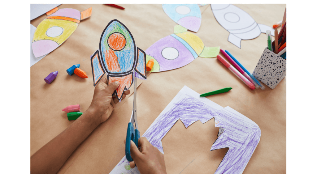 A child cutting out a colorful paper rocket surrounded by crayons and drawings, symbolizing Market Research Money Hacks to launch big ideas creatively.