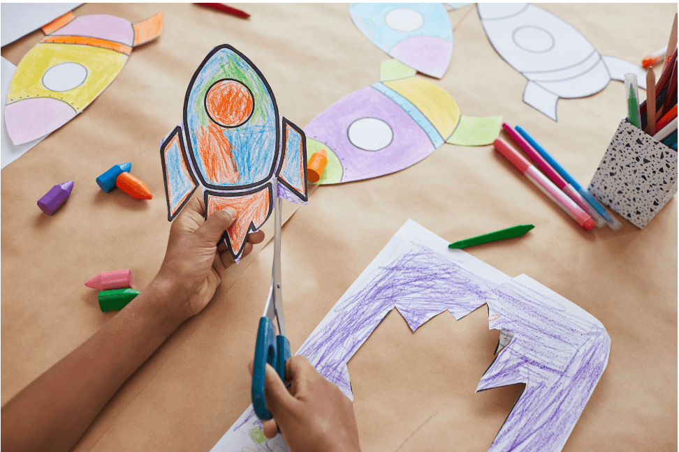 A child cutting out a colorful paper rocket surrounded by crayons and drawings, symbolizing Market Research Money Hacks to launch big ideas creatively.