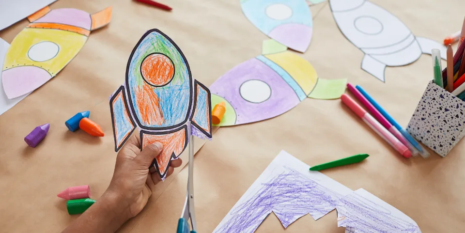 A child’s hands cutting out a colorful, hand-drawn rocket from paper, surrounded by crayons and other rocket drawings on a table, symbolizing creativity and imagination in aiming for big goals